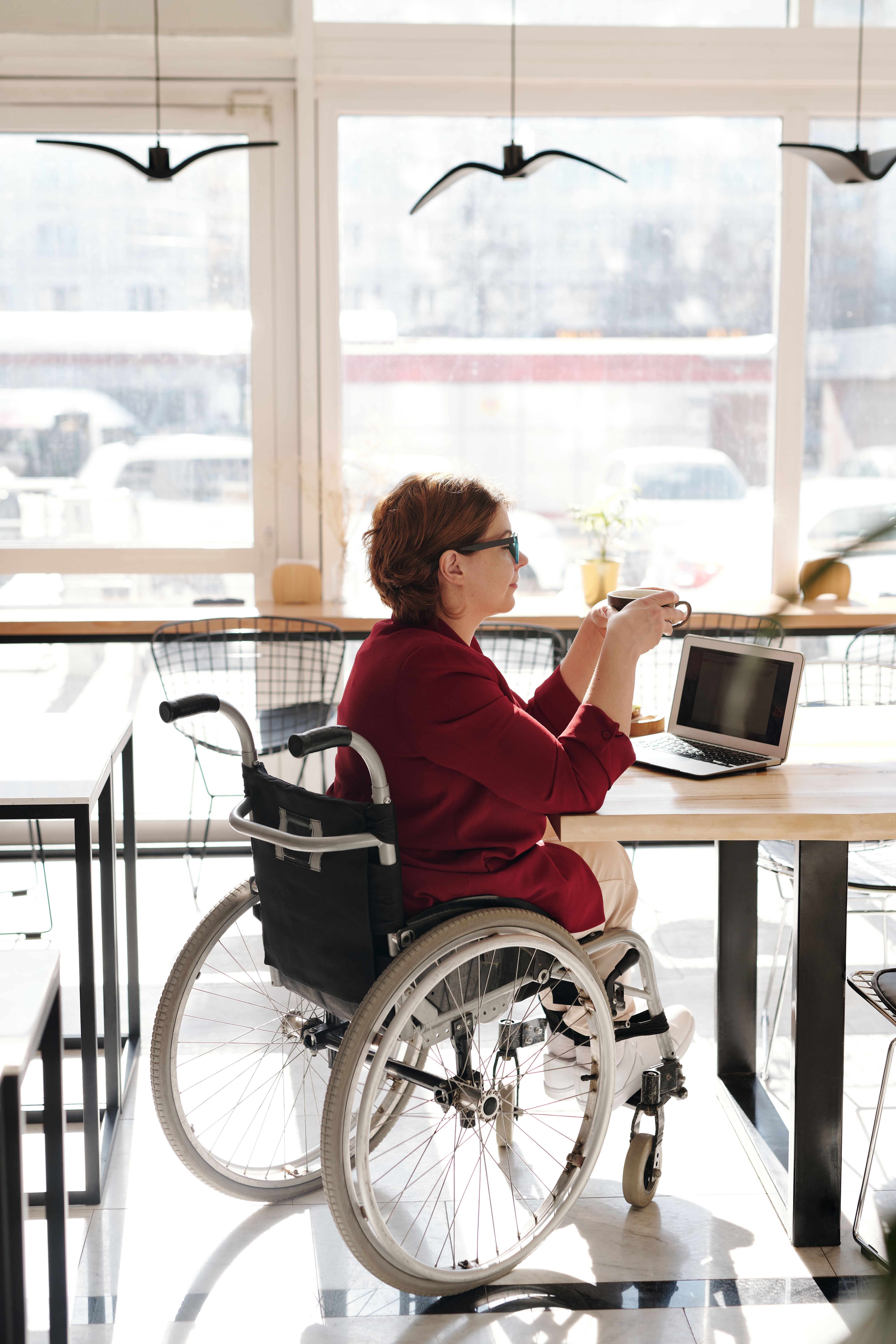 A woman in a wheelchair working on her laptop at a cafe, enjoying her coffee.
