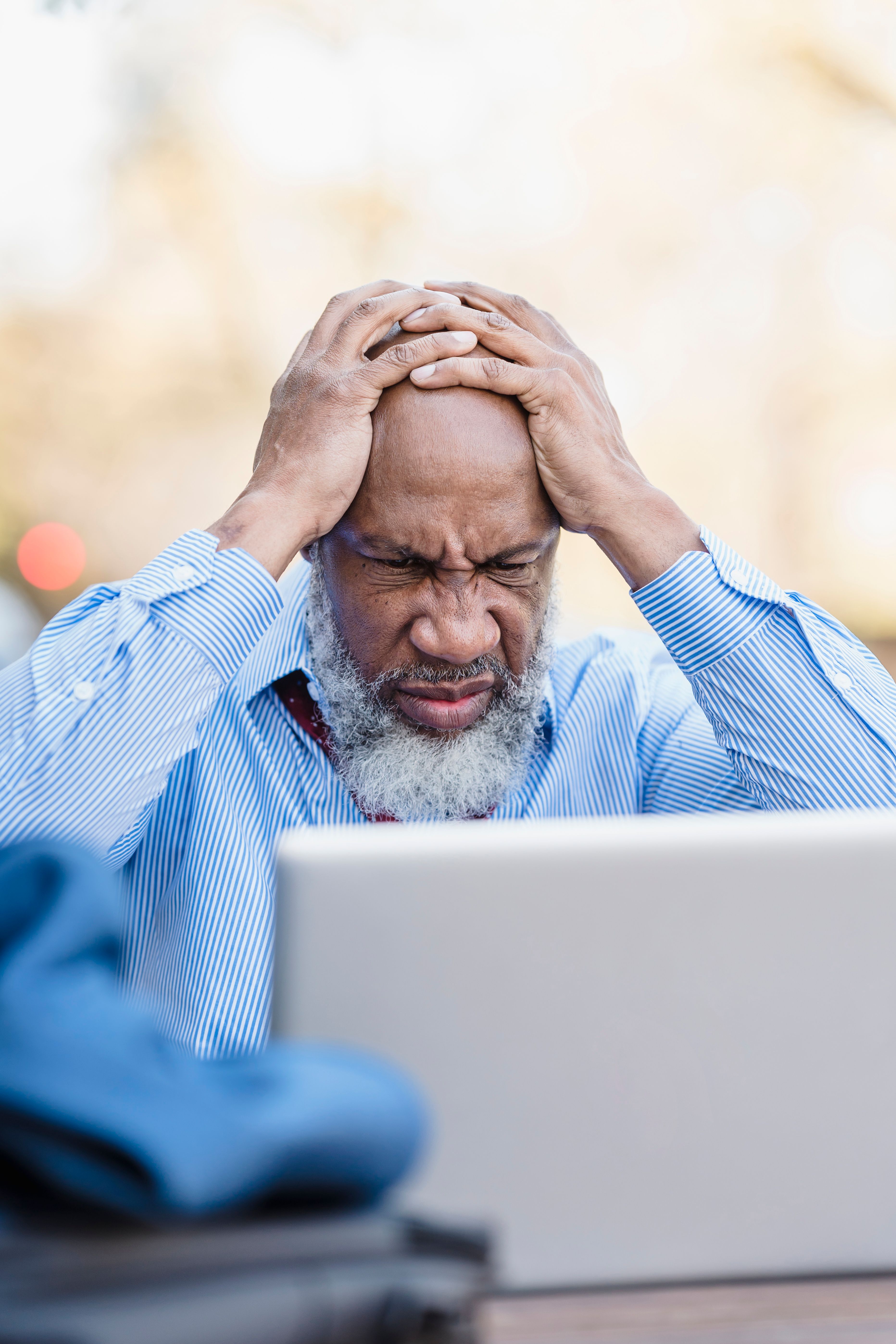 Senior man sitting outdoors with head in hands, appearing stressed while using a laptop.